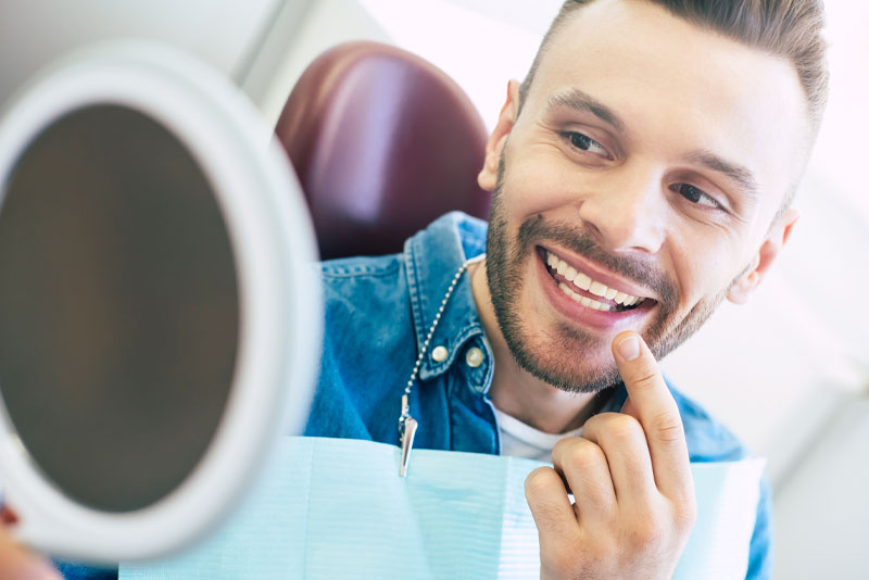 a dental patient smiling at his new smile through a hand held mirror after his crown lengthening procedure.