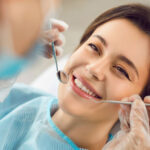 Woman smiling during a dental checkup as a dentist uses a mirror and dental tool to examine her teeth.