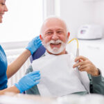 Smiling senior man sitting in a dental chair with a bib on, holding a suction tool, while a dental hygienist stands beside him.