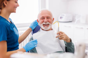 Smiling senior man sitting in a dental chair with a bib on, holding a suction tool, while a dental hygienist stands beside him.